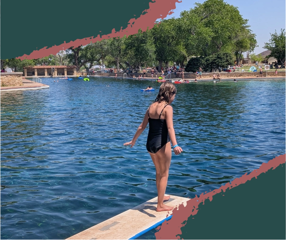Child mid-air jumping into the crystal-clear, spring-fed pool at Balmorhea State Park in Texas, with the San Solomon Courts and pool area visible in the background.