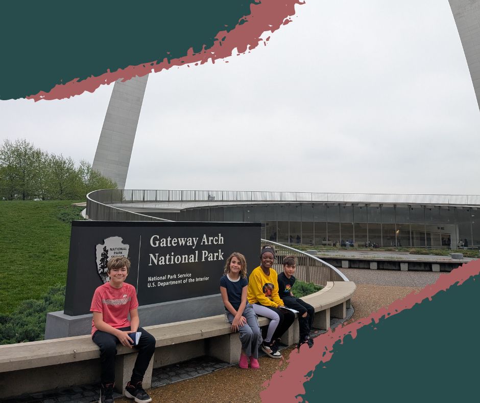 Kids smiling by the Gateway Arch National Park sign with the Gateway Arch rising in the background.